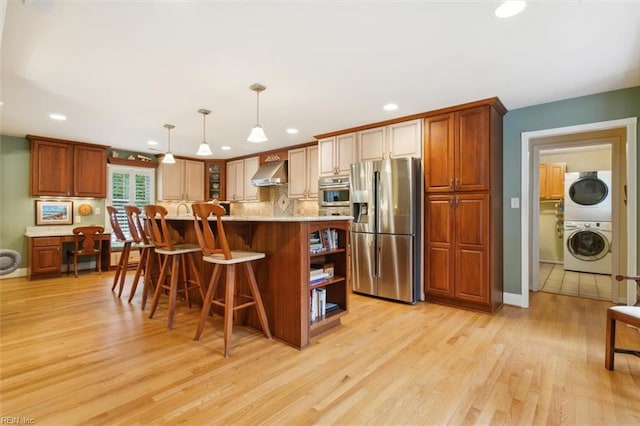 kitchen featuring wall chimney range hood, light hardwood / wood-style flooring, stacked washing maching and dryer, hanging light fixtures, and stainless steel appliances