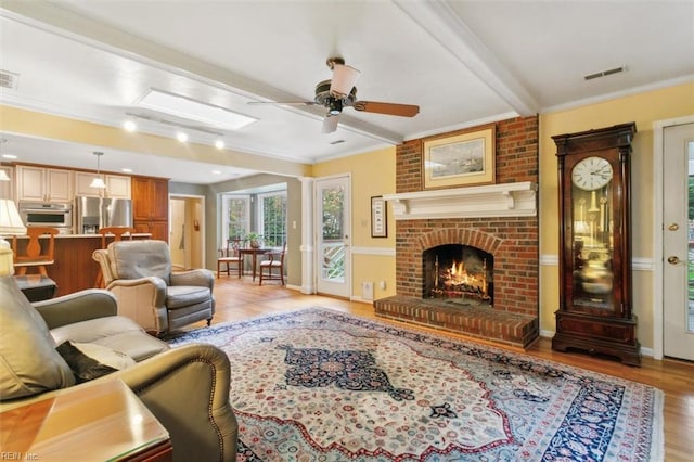 living room with ceiling fan, crown molding, a brick fireplace, beam ceiling, and light hardwood / wood-style flooring