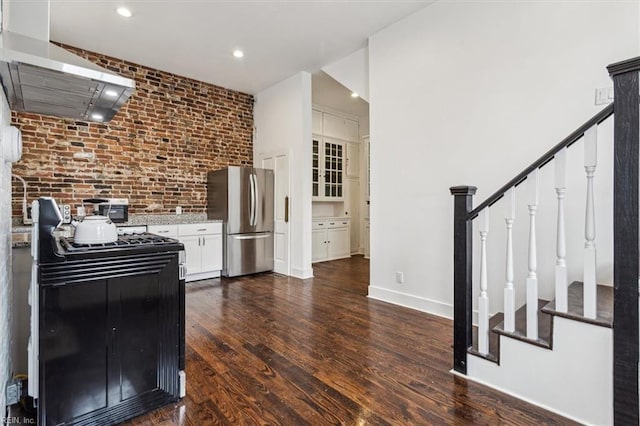 kitchen featuring dark wood-type flooring, gas range, stainless steel fridge, brick wall, and wall chimney range hood