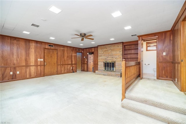 unfurnished living room featuring ceiling fan, a stone fireplace, light colored carpet, and wooden walls