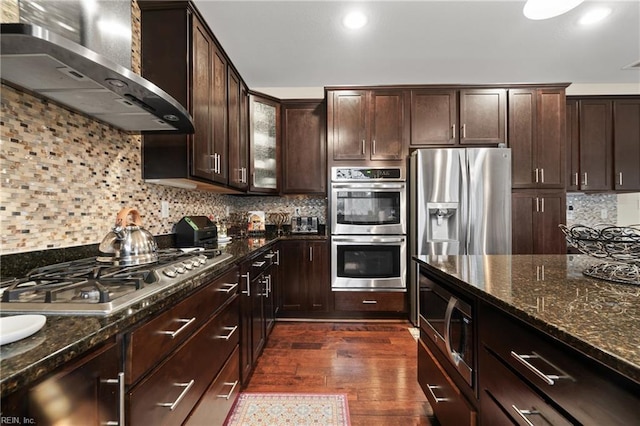 kitchen with stainless steel appliances, wall chimney range hood, dark hardwood / wood-style flooring, and dark stone counters