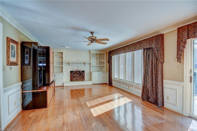 unfurnished living room featuring built in shelves, light wood-type flooring, ornamental molding, ceiling fan, and a fireplace