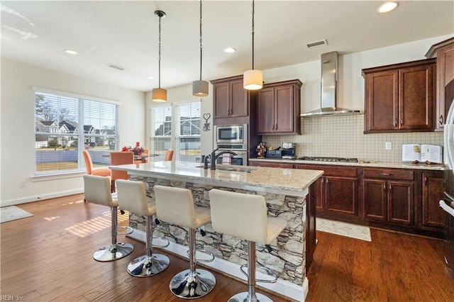 kitchen with wall chimney exhaust hood, light stone counters, appliances with stainless steel finishes, an island with sink, and pendant lighting