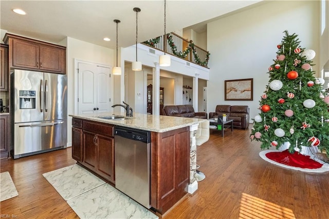 kitchen with sink, hanging light fixtures, stainless steel appliances, light stone countertops, and a center island with sink