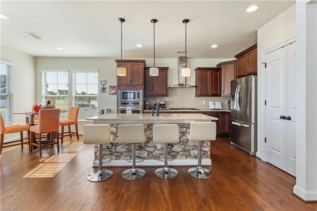 kitchen with dark wood-type flooring, appliances with stainless steel finishes, hanging light fixtures, an island with sink, and wall chimney exhaust hood