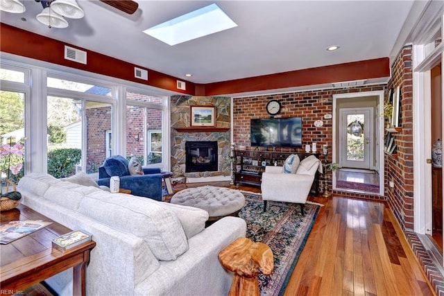 living room featuring hardwood / wood-style flooring, ceiling fan, a skylight, a fireplace, and brick wall