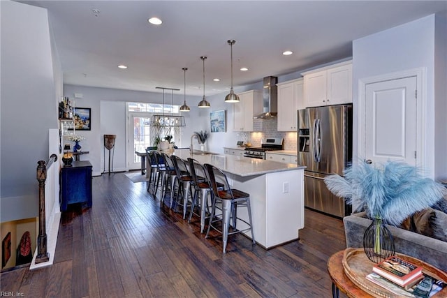 kitchen featuring wall chimney exhaust hood, hanging light fixtures, appliances with stainless steel finishes, a kitchen island with sink, and white cabinets