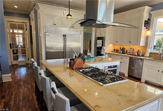 kitchen with appliances with stainless steel finishes, hanging light fixtures, island exhaust hood, light stone counters, and a center island with sink