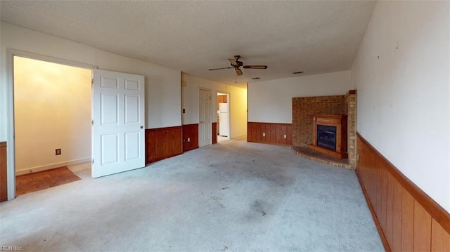 unfurnished living room featuring light carpet, wooden walls, a wainscoted wall, ceiling fan, and a brick fireplace