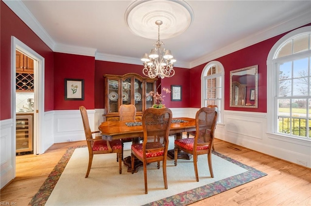 dining area with wine cooler, an inviting chandelier, light wood-style floors, ornamental molding, and wainscoting