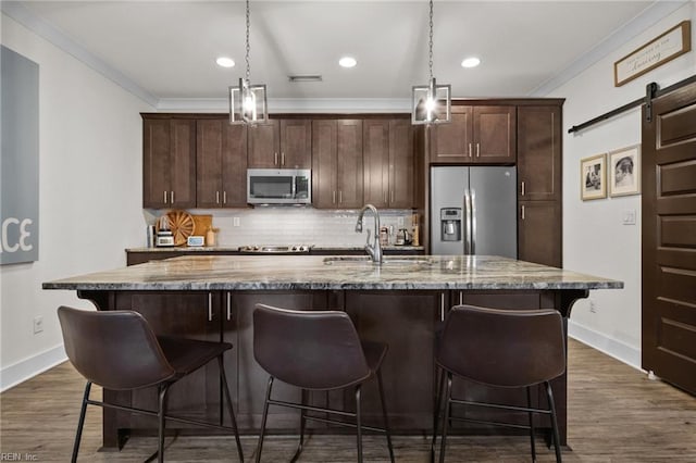 kitchen with ornamental molding, light stone counters, stainless steel appliances, a barn door, and a center island with sink