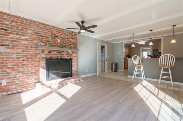 unfurnished living room featuring ceiling fan, brick wall, a brick fireplace, beamed ceiling, and light wood-type flooring