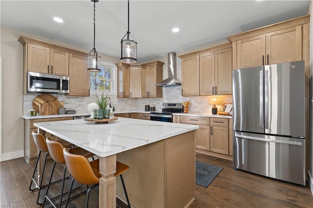 kitchen with wall chimney range hood, appliances with stainless steel finishes, hanging light fixtures, a kitchen island, and light brown cabinetry