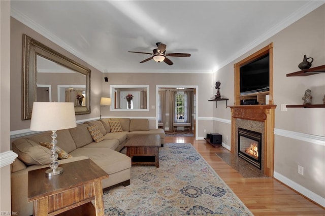 living area featuring a fireplace with flush hearth, a ceiling fan, baseboards, light wood-type flooring, and crown molding