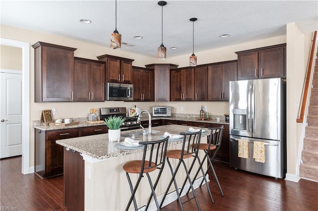 kitchen with light stone counters, dark wood-type flooring, appliances with stainless steel finishes, an island with sink, and pendant lighting