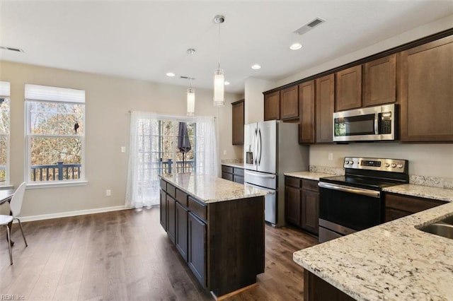 kitchen with light stone counters, dark brown cabinetry, stainless steel appliances, hanging light fixtures, and a center island