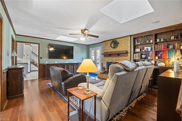 living room featuring light wood-type flooring, a brick fireplace, ornamental molding, ceiling fan, and a skylight