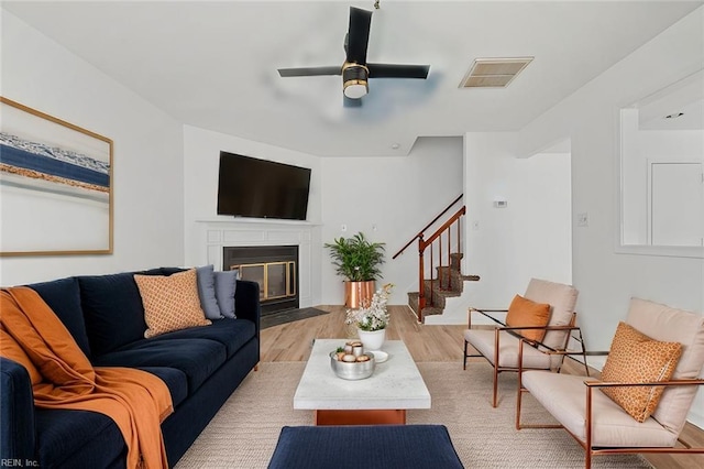 living area with light wood-type flooring, stairs, a fireplace with flush hearth, a ceiling fan, and visible vents