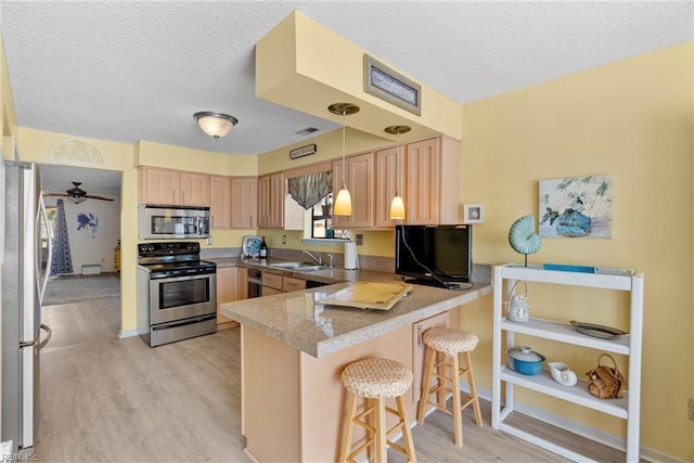 kitchen with light brown cabinetry, appliances with stainless steel finishes, a sink, light wood-type flooring, and a peninsula