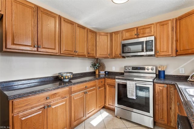 kitchen with light tile patterned floors, appliances with stainless steel finishes, brown cabinets, and dark stone counters