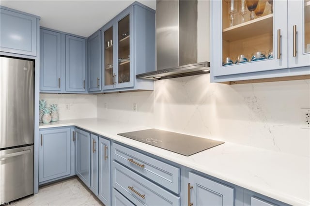 kitchen with black electric stovetop, light countertops, glass insert cabinets, freestanding refrigerator, and wall chimney range hood