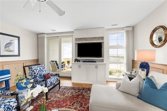 living room with a wainscoted wall, ceiling fan, visible vents, and light wood-style floors