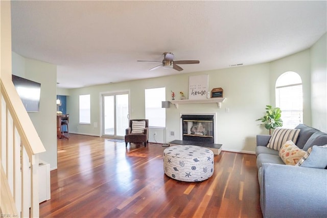 living room featuring ceiling fan, visible vents, dark wood finished floors, baseboards, and a glass covered fireplace