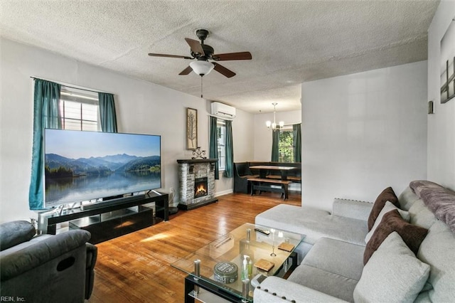 living room featuring a wall mounted AC, a stone fireplace, a textured ceiling, wood finished floors, and ceiling fan with notable chandelier