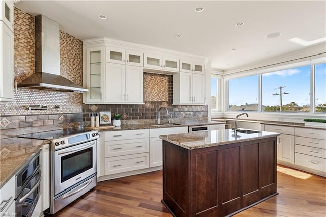 kitchen with wall chimney exhaust hood, a center island with sink, glass insert cabinets, and stainless steel appliances