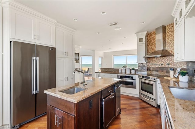 kitchen featuring wall chimney exhaust hood, glass insert cabinets, appliances with stainless steel finishes, a kitchen island with sink, and a sink