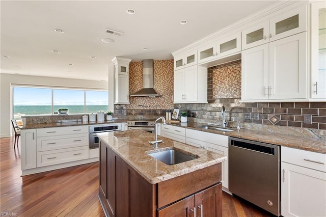 kitchen featuring a center island with sink, appliances with stainless steel finishes, a water view, wall chimney range hood, and a sink