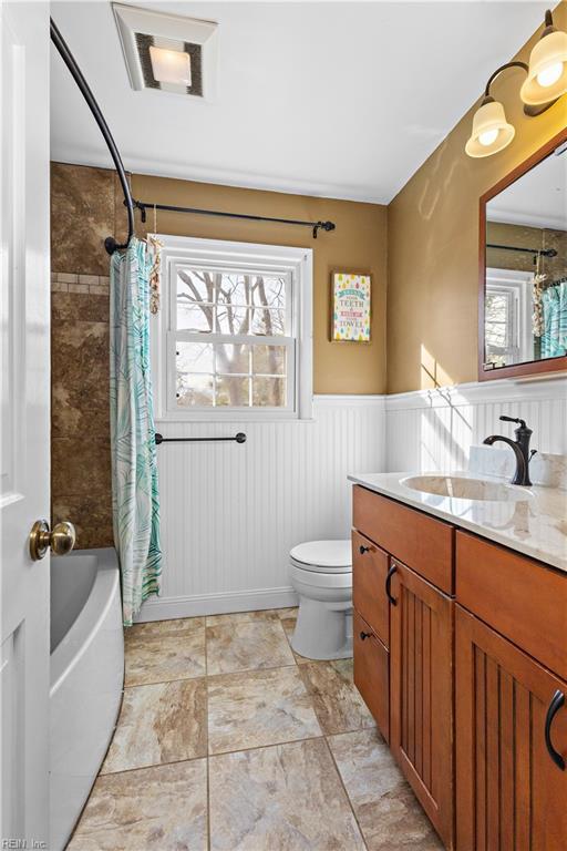 full bath with a wainscoted wall, shower / bathtub combination with curtain, toilet, and vanity