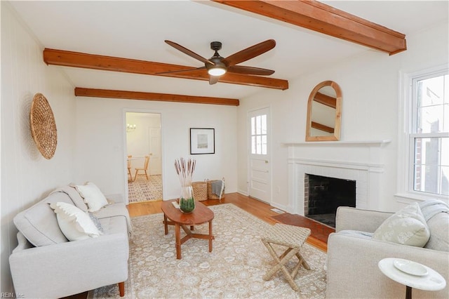 living room featuring a ceiling fan, a brick fireplace, beam ceiling, and light wood finished floors