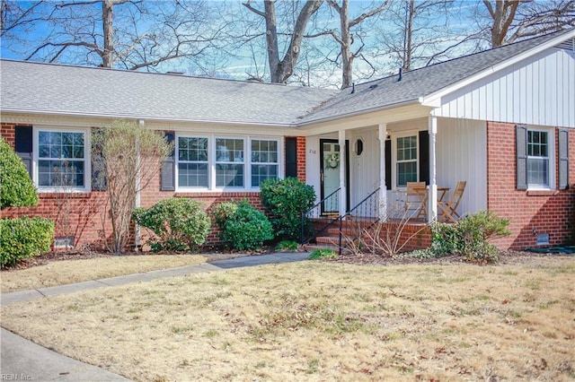 doorway to property featuring a porch, brick siding, a lawn, and roof with shingles