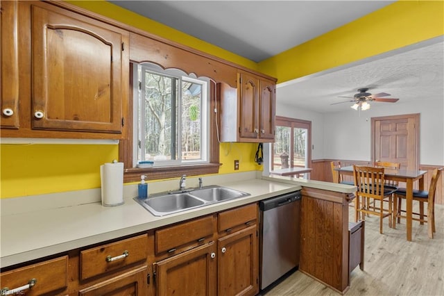 kitchen with a wainscoted wall, brown cabinets, light countertops, stainless steel dishwasher, and a sink