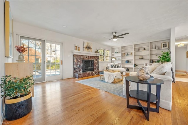 living room with built in features, a high end fireplace, ceiling fan, and light wood finished floors