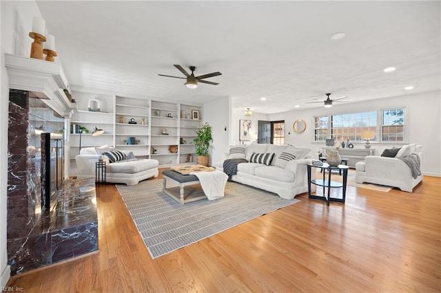 living room featuring built in shelves, light wood-style flooring, recessed lighting, a premium fireplace, and a ceiling fan