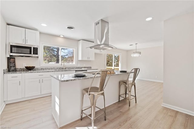 kitchen featuring island range hood, a kitchen island, white cabinetry, stainless steel microwave, and decorative light fixtures