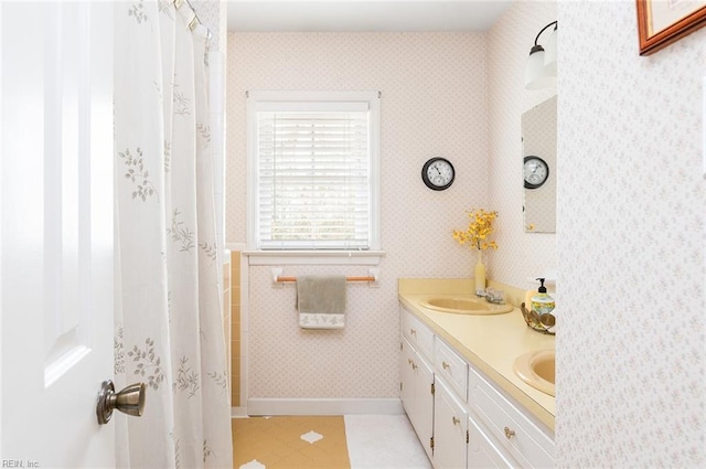 full bathroom with baseboards, double vanity, a sink, and wallpapered walls