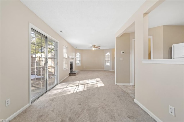 empty room with light carpet, baseboards, a fireplace with raised hearth, and a ceiling fan