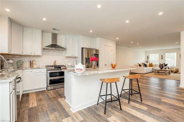 kitchen featuring white cabinets, open floor plan, appliances with stainless steel finishes, a center island, and wall chimney exhaust hood