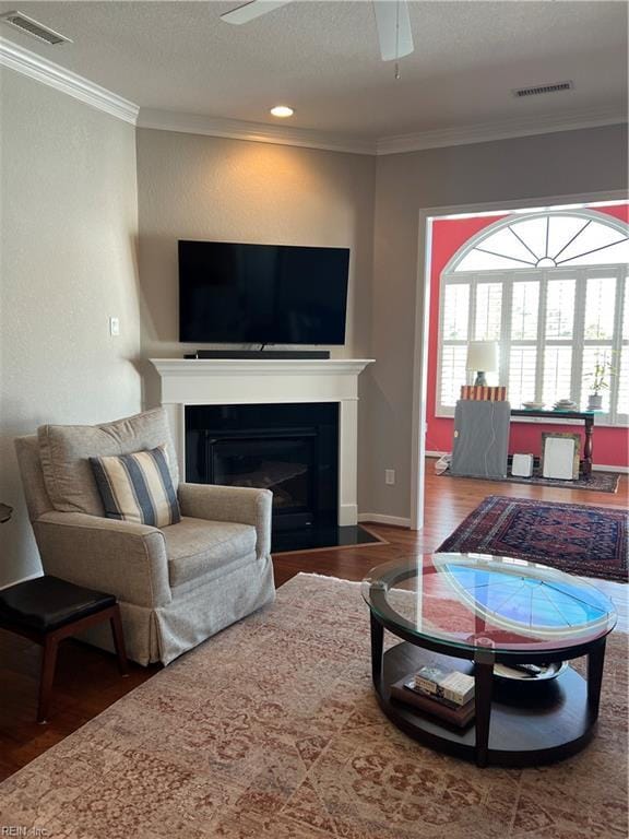 living room with a fireplace with flush hearth, a ceiling fan, wood finished floors, visible vents, and crown molding