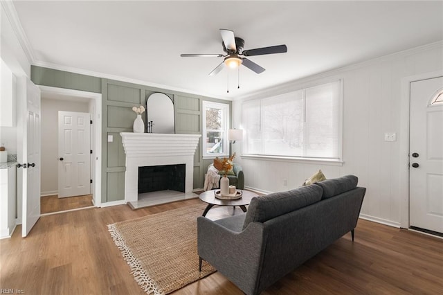 living area with baseboards, a fireplace with raised hearth, ceiling fan, wood finished floors, and crown molding