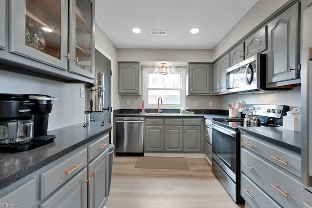 kitchen with gray cabinetry, stainless steel appliances, a sink, dark countertops, and glass insert cabinets