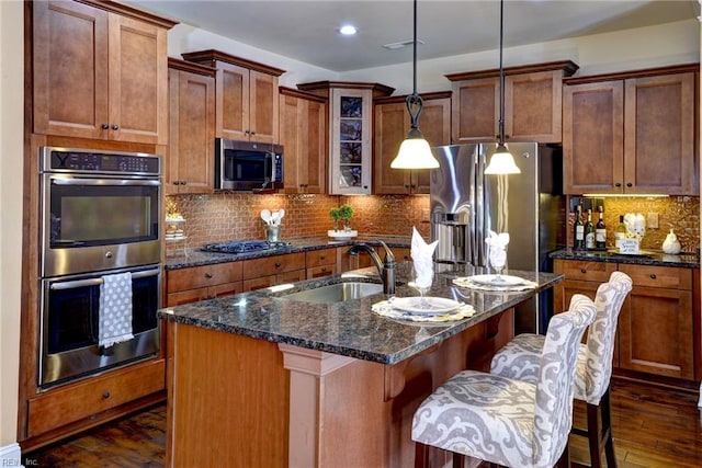 kitchen featuring an island with sink, brown cabinets, stainless steel appliances, and hanging light fixtures