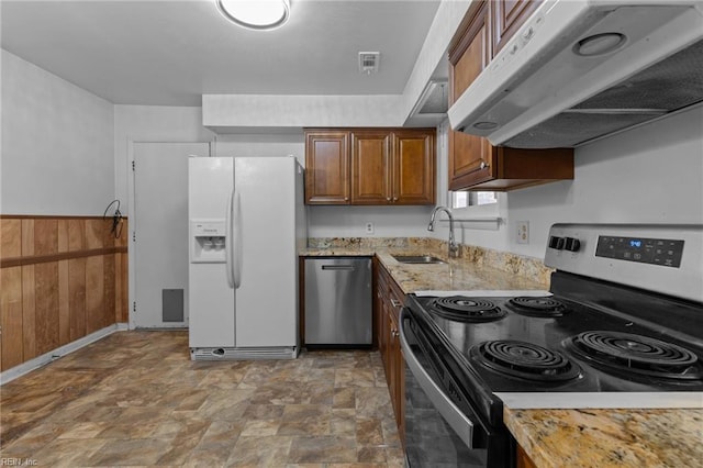 kitchen featuring appliances with stainless steel finishes, light stone countertops, wood walls, under cabinet range hood, and a sink