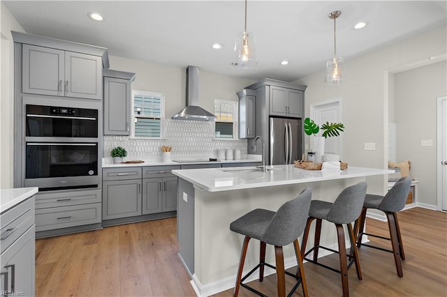 kitchen featuring light countertops, double oven, freestanding refrigerator, a kitchen island with sink, and wall chimney range hood