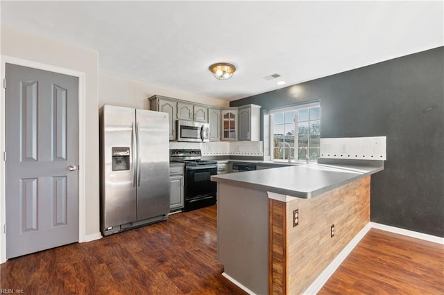 kitchen featuring glass insert cabinets, appliances with stainless steel finishes, dark wood-style flooring, a peninsula, and gray cabinets