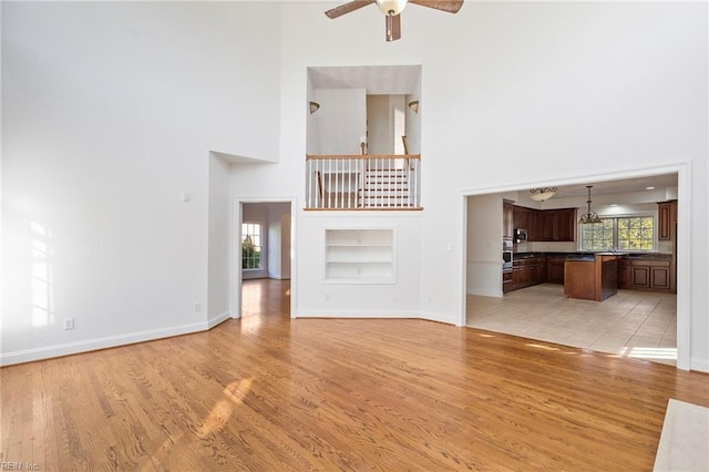 unfurnished living room featuring a towering ceiling, ceiling fan, light wood-style flooring, and baseboards