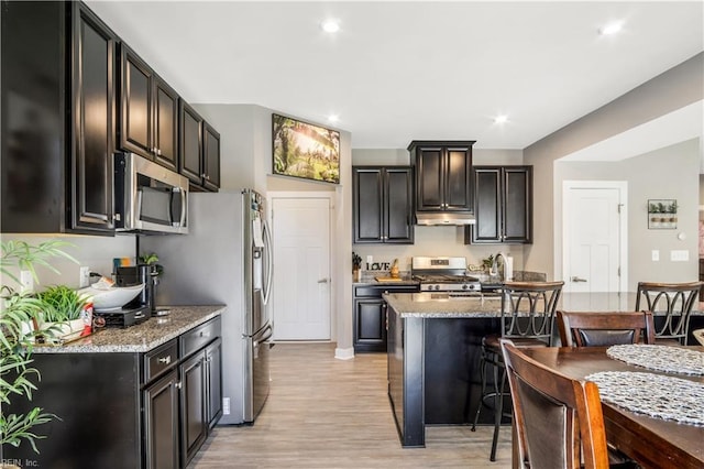 kitchen with light wood-style flooring, appliances with stainless steel finishes, light stone counters, under cabinet range hood, and a kitchen bar